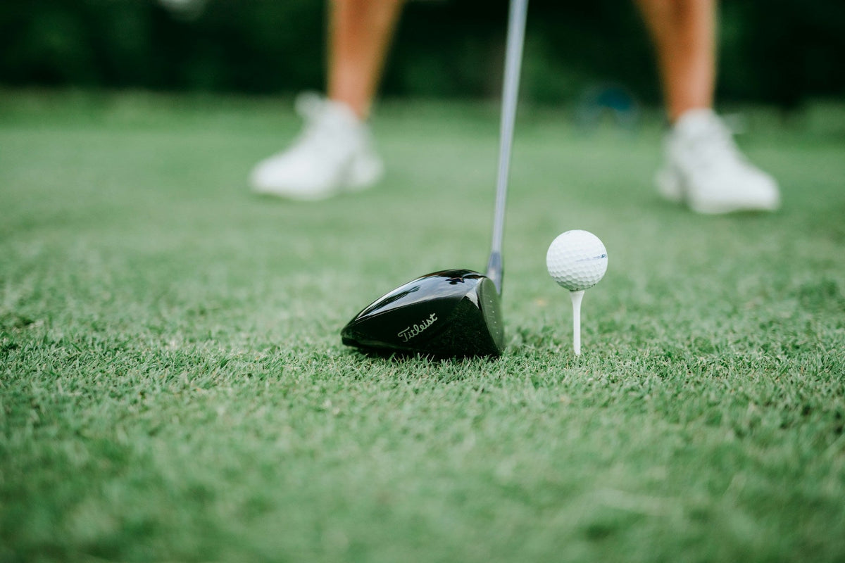 close look at the head of a driver golf club about to hit a ball with the feet of the golf player in the background over a green grass