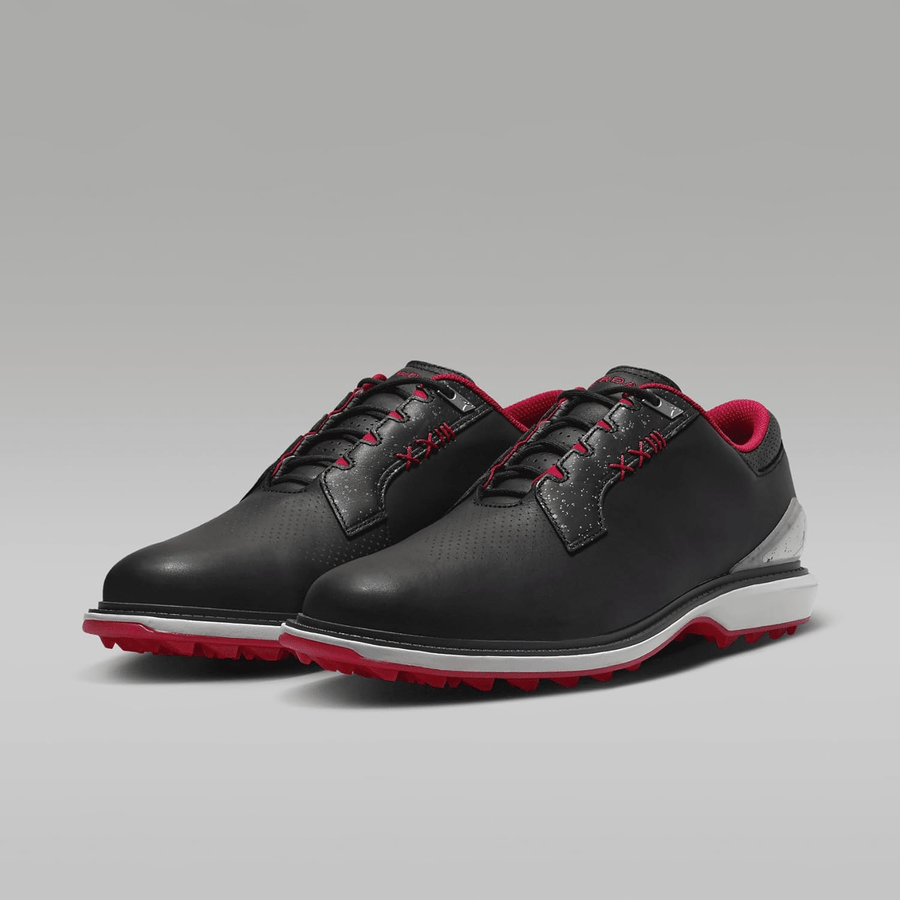 The Jordan ADG 5 Golf Shoes - Black feature red and white accents, red laces and soles, a sleek design with perforated details, and a signature rubber traction pattern, shown against a plain grey background.