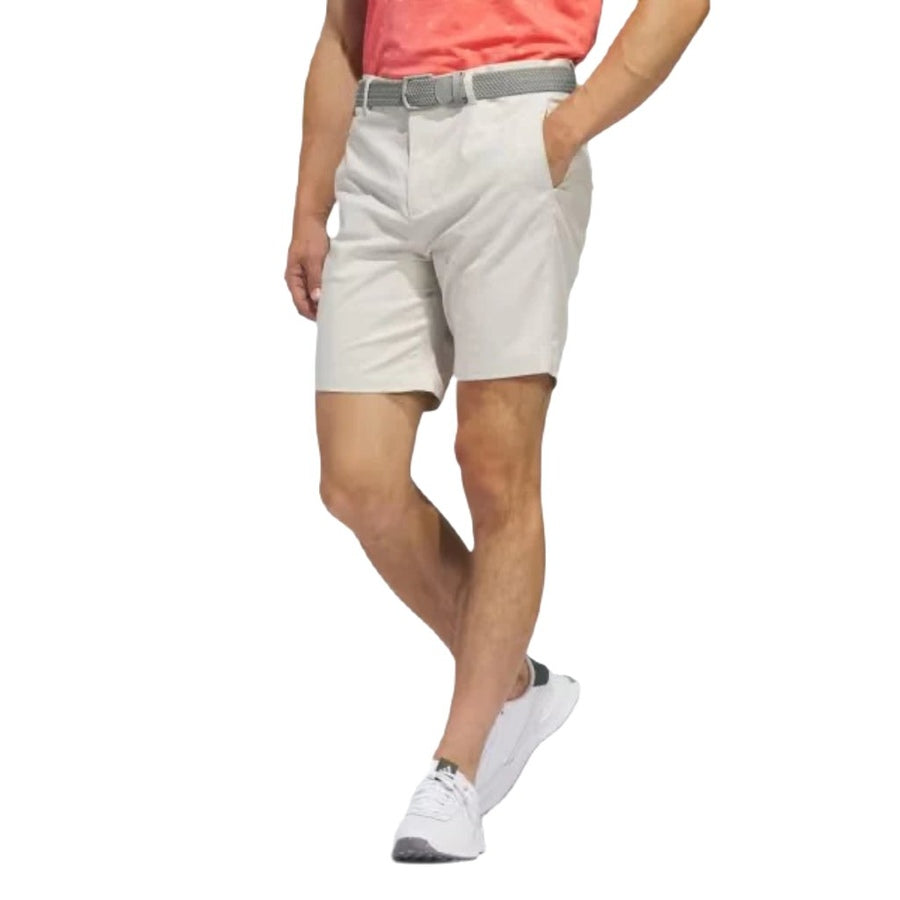 A person in a coral t-shirt, gray belt, white adidas sneakers, and light beige Adidas Go-To Five-Pocket Golf Shorts stands with hands in pockets against a white background.
