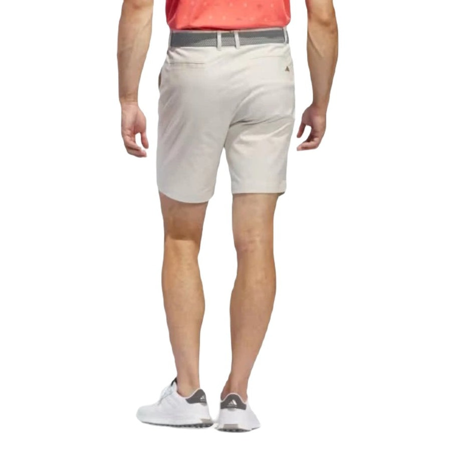 A person wearing a red shirt, gray belt, white sneakers, and Adidas Go-To Five-Pocket Golf Shorts in beige is seen from the back, showcasing the versatility of their outfit against a white background.