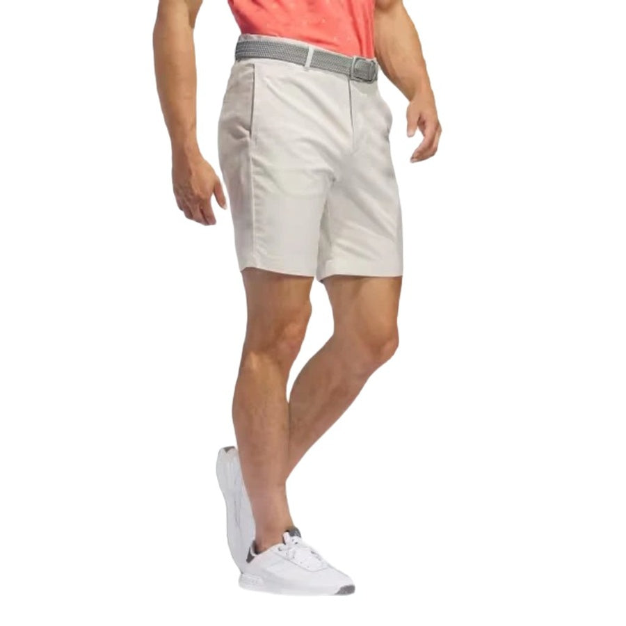A man wearing Adidas Go-To Five-Pocket Golf Shorts, a coral t-shirt, a gray woven belt, and white adidas sneakers stands with one leg bent against a white background.