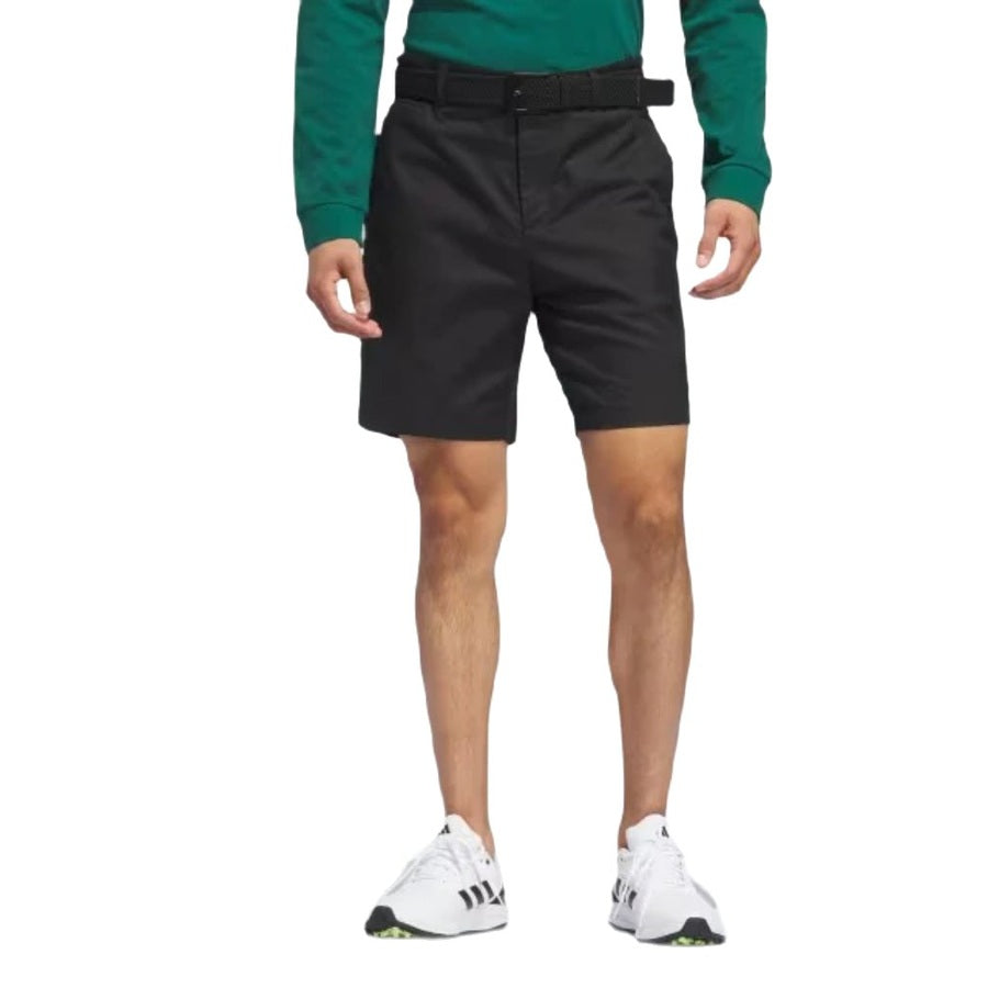 A person stands against a plain background wearing a green long-sleeve shirt, Adidas Go-To Five-Pocket Golf Shorts with a belt, and white adidas athletic shoes with black stripes. Their head is not visible in the image.