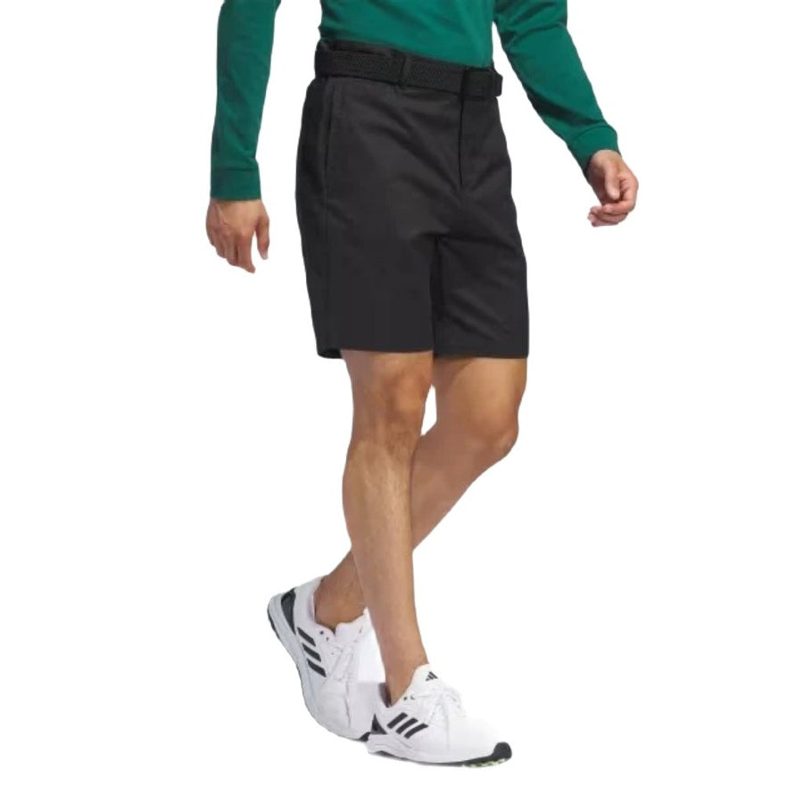 A person is walking, wearing a green long-sleeve shirt, white athletic shoes, and versatile Adidas Go-To Five-Pocket Golf Shorts with a belt. Only the lower half of the body is visible against a white background.
