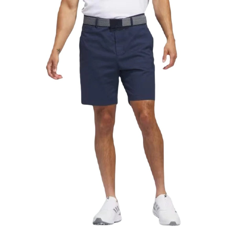 Someone stands against a plain white background wearing Adidas Go-To Five-Pocket Golf Shorts, paired with a white belt, white athletic shoes, and a white shirt. The image is cropped at the waist and neck.