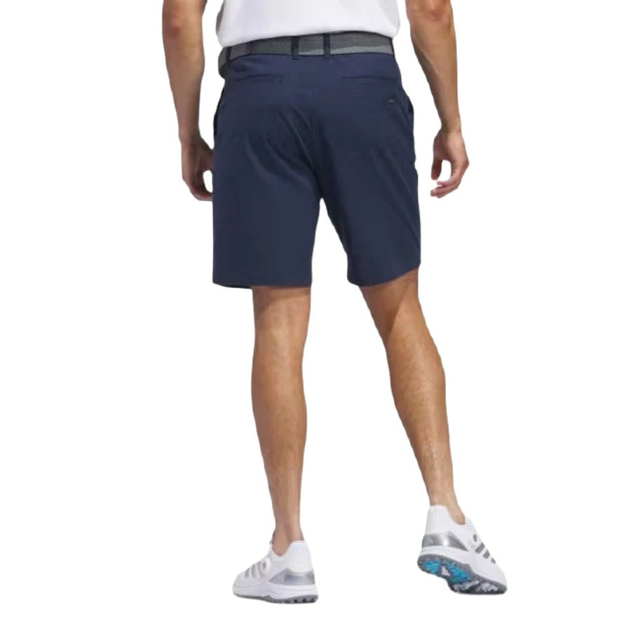 A man in a white shirt, Adidas Go-To Five-Pocket Golf Shorts in dark blue, and white athletic shoes stands with his back to the camera against a plain white background, highlighting a versatile sporty style.