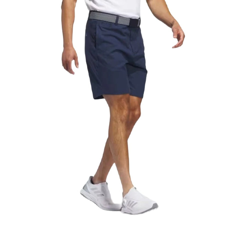 A man wearing a white shirt, Adidas Go-To Five-Pocket Golf Shorts in navy with a gray belt, and white adidas athletic shoes walks against a white background. His face is not visible.