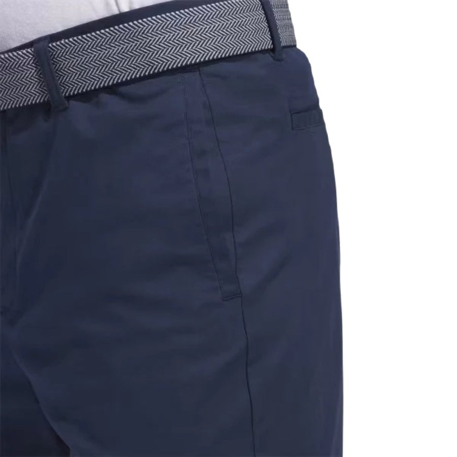 A close-up of a person wearing Adidas Go-To Five-Pocket Golf Shorts in navy blue, paired with a gray and white patterned belt and a tucked-in white shirt.
