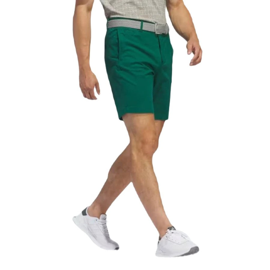 A man in a light gray shirt, Adidas Go-To Five-Pocket Golf Shorts in green with a belt, and white athletic shoes walks against a white background.