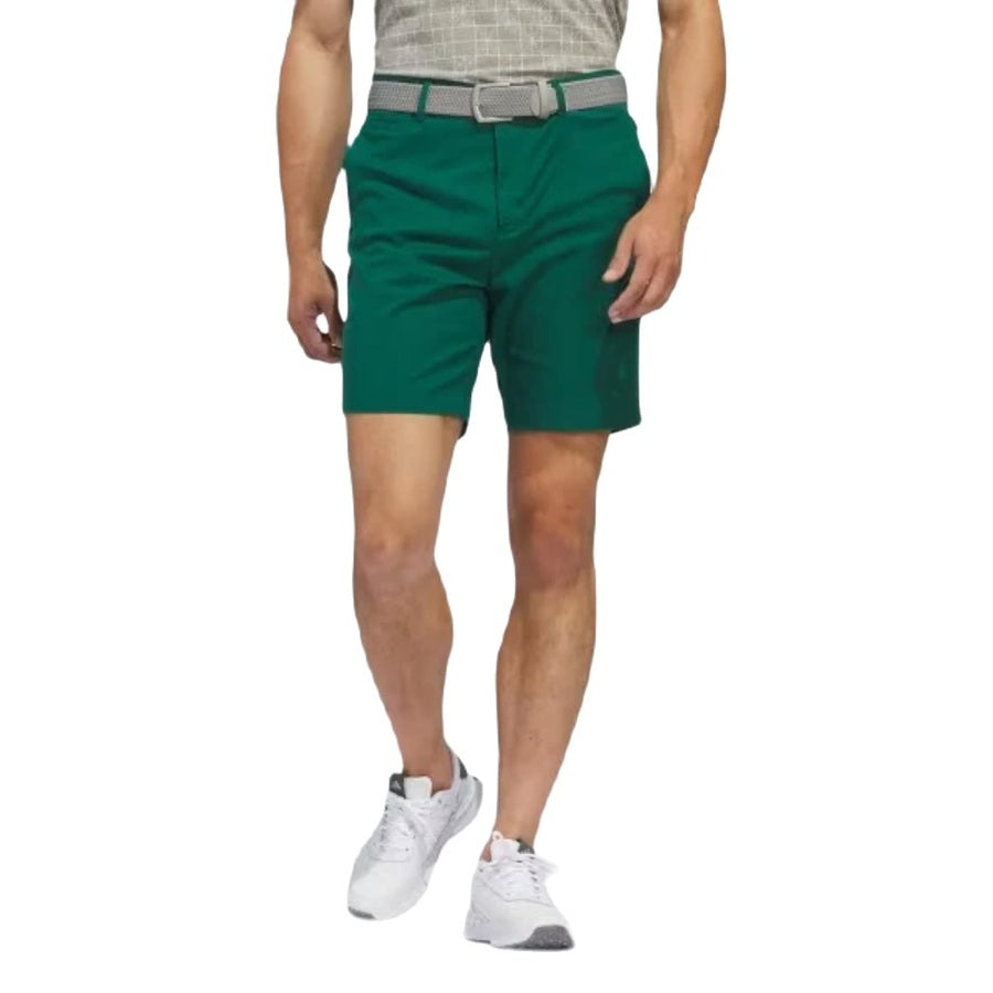 A person stands against a white background wearing a short-sleeve shirt, a gray belt, white athletic shoes, and versatile green Adidas Go-To Five-Pocket Golf Shorts. The persons head is not visible in the image.