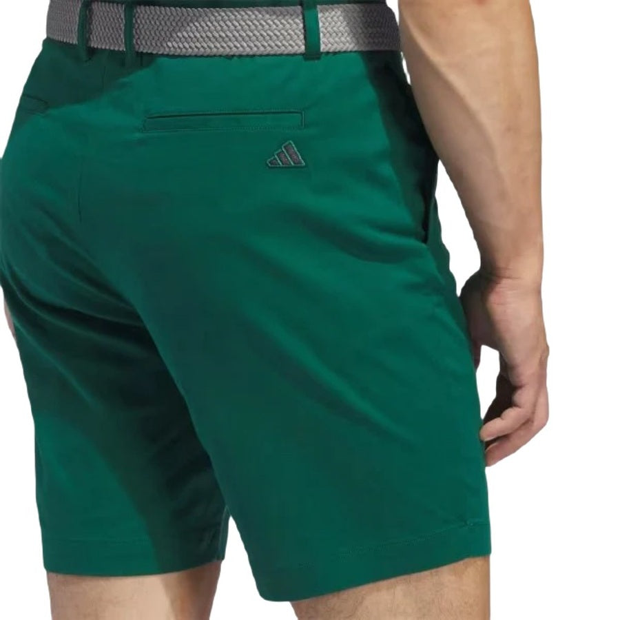 A person from behind wears green Adidas Go-To Five-Pocket Golf Shorts with a gray belt. The shorts display a small Adidas logo above the back pocket.