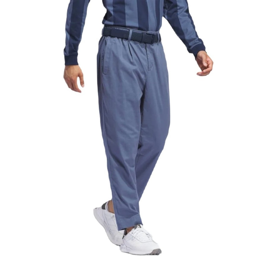 Une personne porte un pantalon adidas Go-To Versatile bleu, en matières recyclées, une chemise rayée bleu et marine à manches longues, une ceinture noire et des baskets blanches, debout, un pied en avant, sur fond blanc uni.