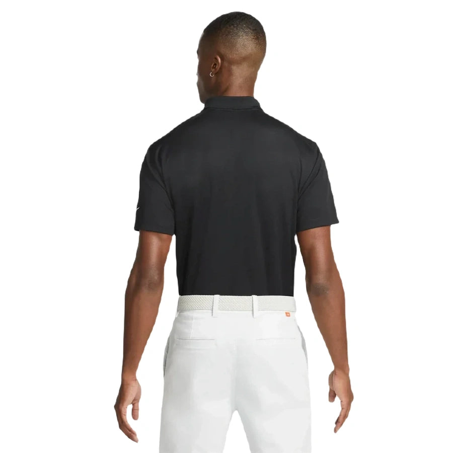 A man stands facing away, wearing the Nike Dri-FIT Victory Men's Golf Shirt with a course/event logo, tucked into white pants with a white belt. The background is plain white.