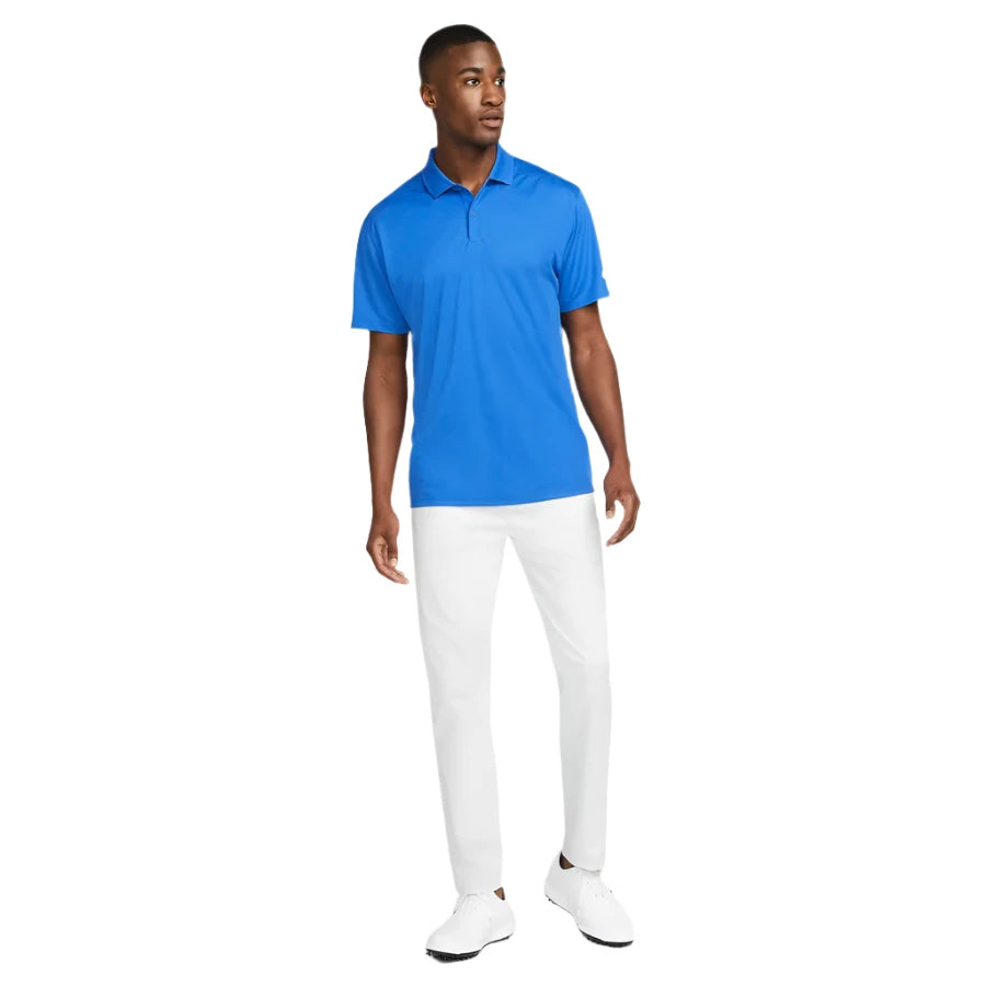 A man stands slightly turned, wearing a bright blue Nike Dri-FIT Victory Men’s Golf Shirt with a course/event logo (final sale), white pants, and white shoes against a plain white background.