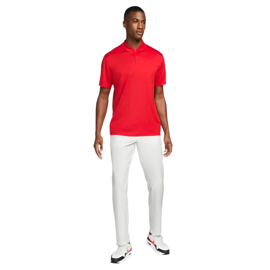 A man stands slightly to the side against a white background, wearing the Nike Dri-FIT Victory Men's Golf Shirt - Course/Event Logo in bright red, paired with white pants and white sneakers featuring red and black accents.
