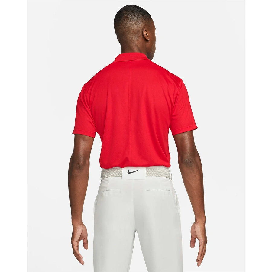 A man faces away, wearing the Nike Dri-FIT Victory Men's Golf Shirt with a course/event logo and white pants with a white belt. The plain background is white.
