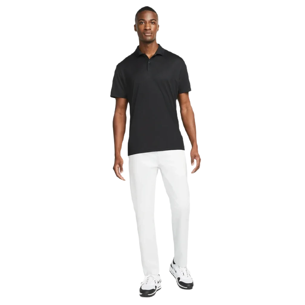 A man stands against a white background wearing the Nike Dri-FIT Victory Men's Golf Shirt with a course/event logo, paired with white pants and white sneakers with black accents.
