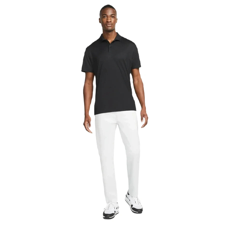 A man stands against a white background wearing the Nike Dri-FIT Victory Men's Golf Shirt with a course/event logo, paired with white pants and white sneakers with black accents.