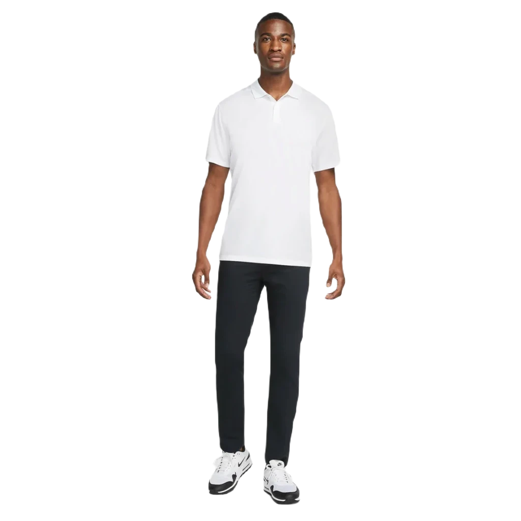 A man stands facing forward in a white Nike Dri-FIT Victory Men's Golf Shirt - Course/Event Logo (Final Sale), black pants, and white sneakers with black accents. The background is plain white.