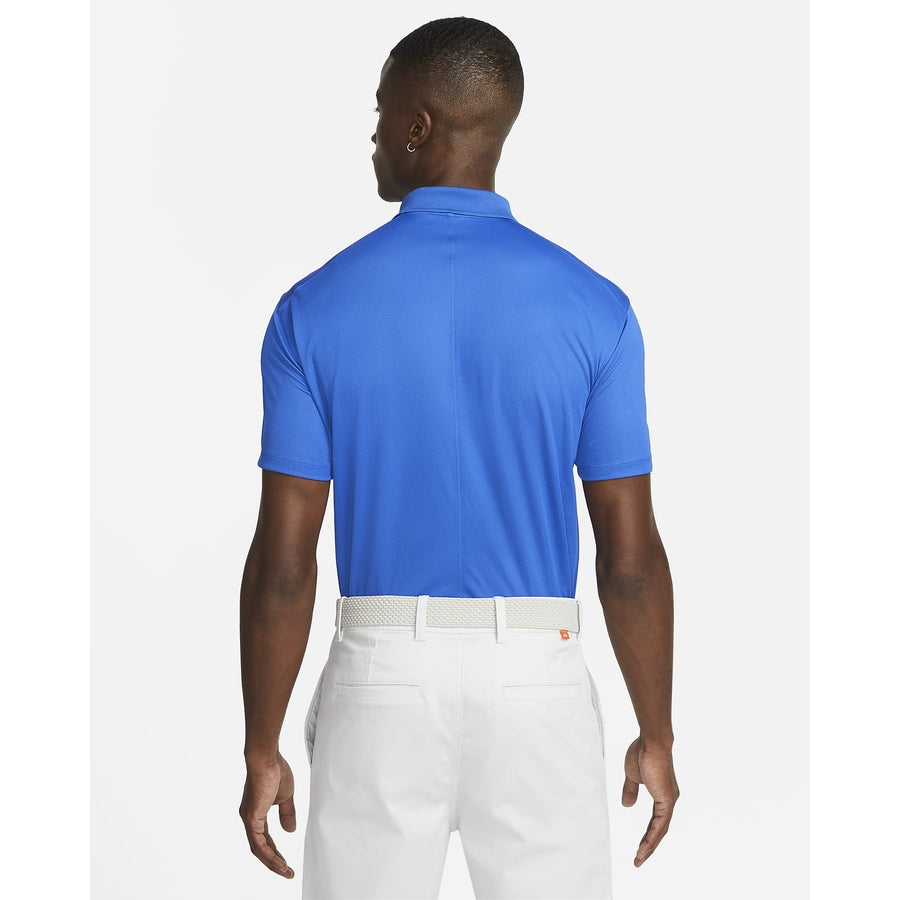 A man is seen from behind wearing a Nike Dri-FIT Victory Men's Golf Shirt with a course/event logo, tucked into white pants and paired with a white belt, standing against a plain white background.