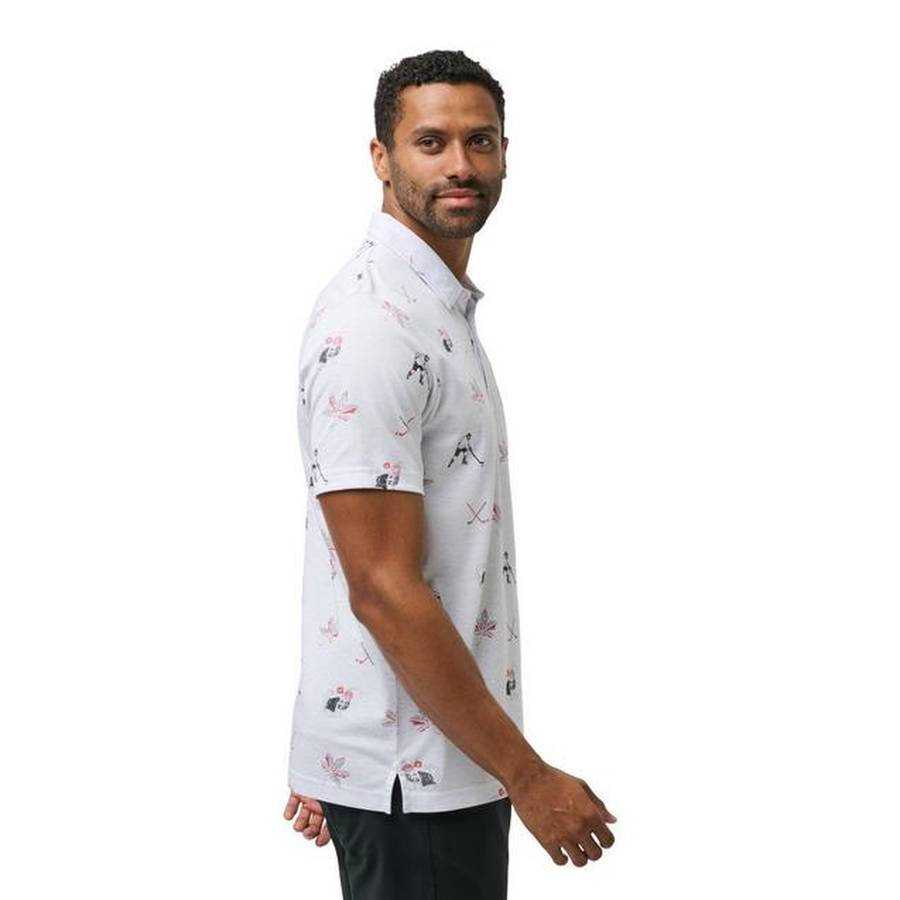 A man with short curly hair and a beard wears the Travis Mathew Bold as Brass Golf Shirt, a light-colored, short-sleeve polo with a subtle print, facing sideways and looking at the camera against a plain white background.