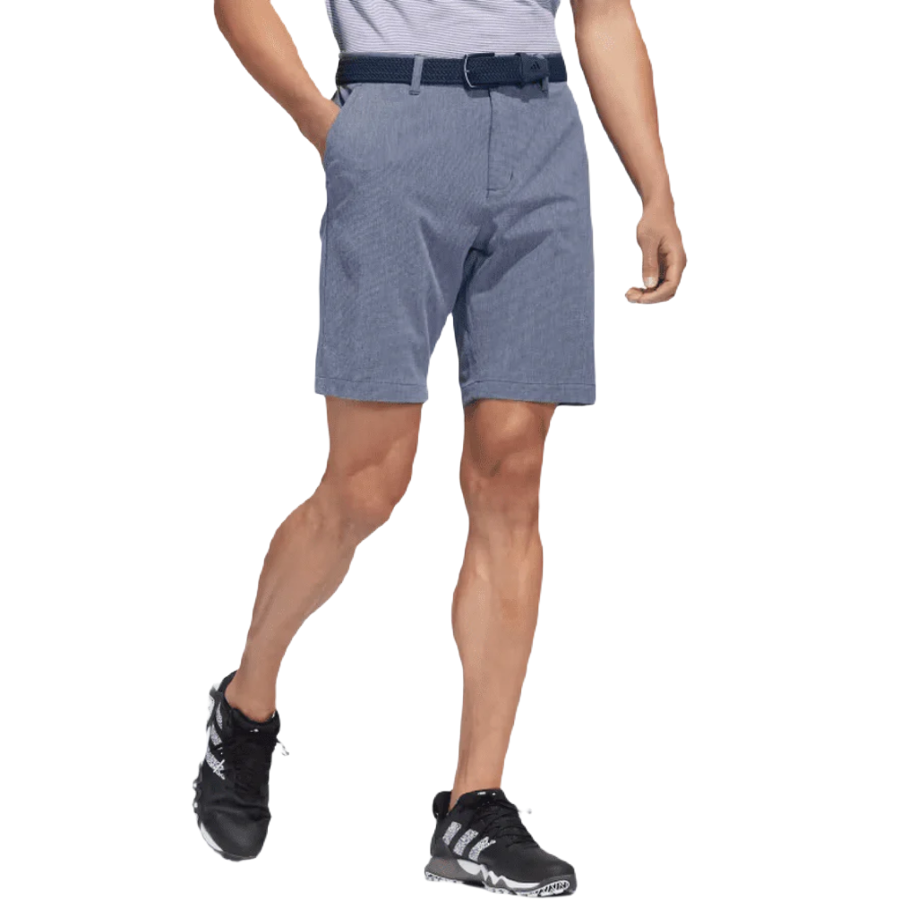 A person stands with hands in pockets against a white background, wearing Adidas Crosshatch Shorts in blue, paired with a light-striped tucked-in shirt and black athletic sneakers.