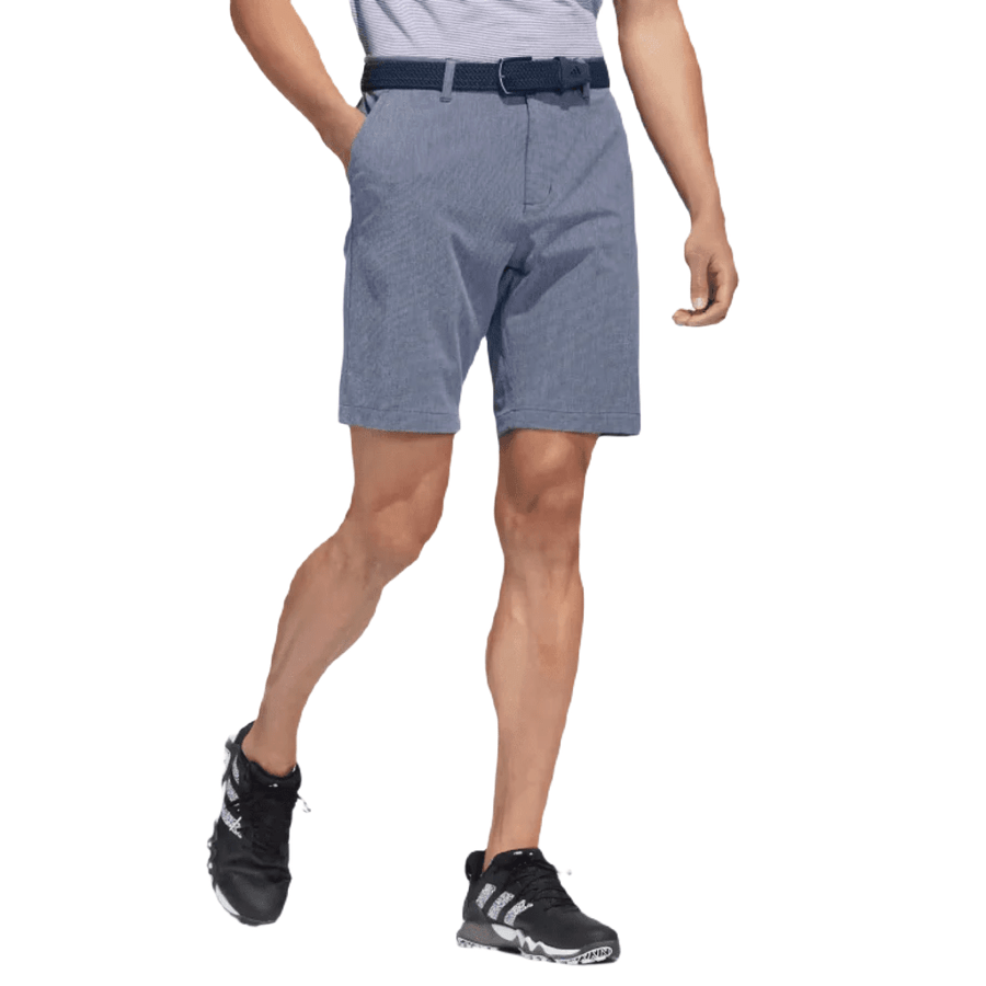 A person stands with hands in pockets against a white background, wearing Adidas Crosshatch Shorts in blue, paired with a light-striped tucked-in shirt and black athletic sneakers.