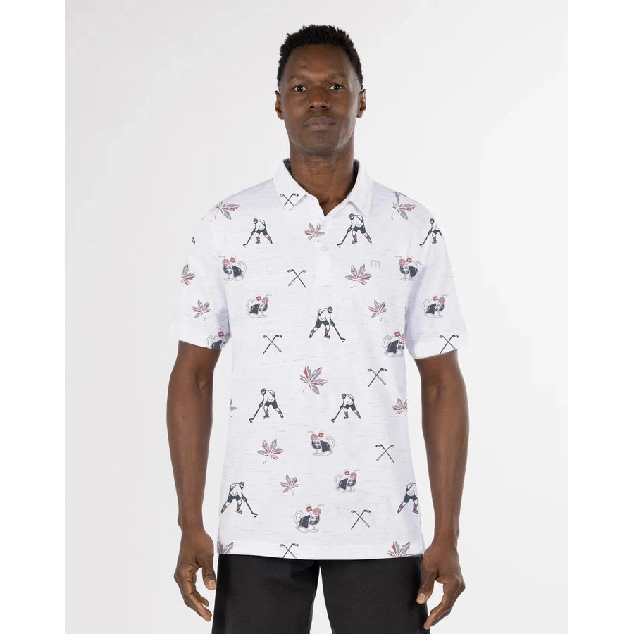 A man faces forward, arms relaxed, wearing the Travis Mathew Bold as Brass Golf Shirt—a white short-sleeve polo with an all-over print of golfers, golf clubs, and red maple leaves against a plain white background.
