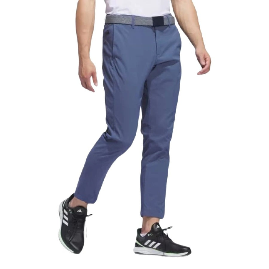 A person wears blue Adidas Ultimate365 Chino Pants made from recycled materials, a woven belt, and black Adidas sneakers with white stripes, shown from mid-torso down against a white background.