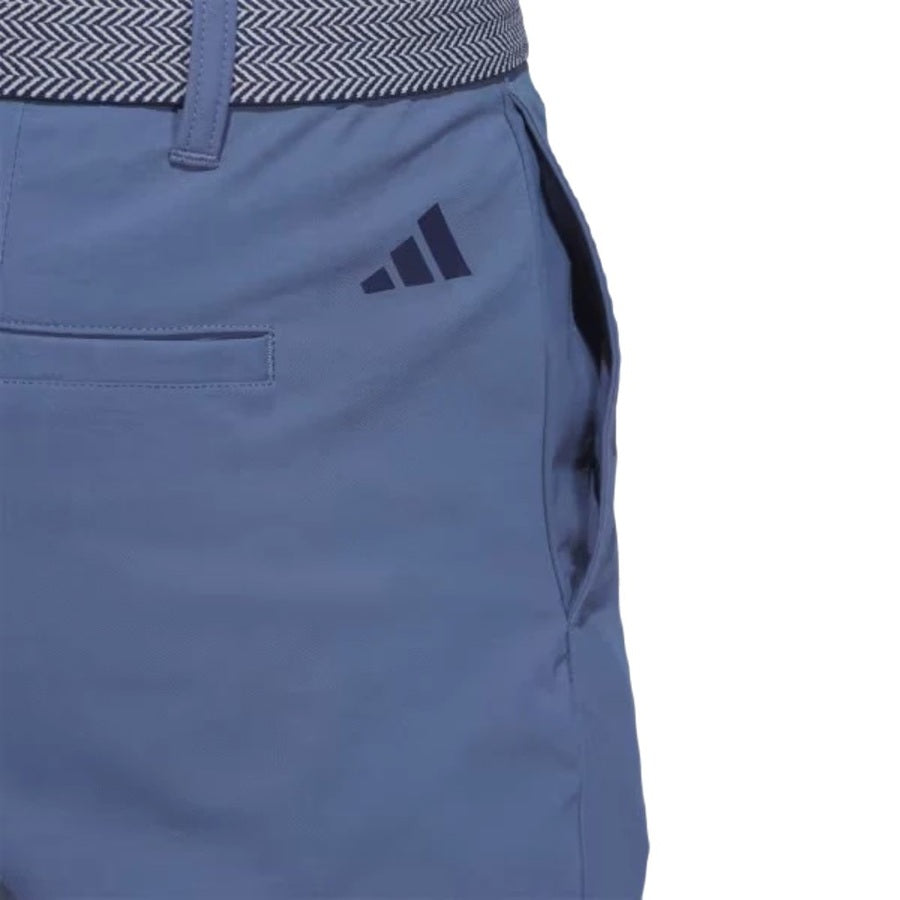 Close-up of a person wearing blue Adidas Ultimate365 Chino Pants featuring a zigzag-patterned waistband and a navy Adidas logo near the front pocket, made from recycled materials.