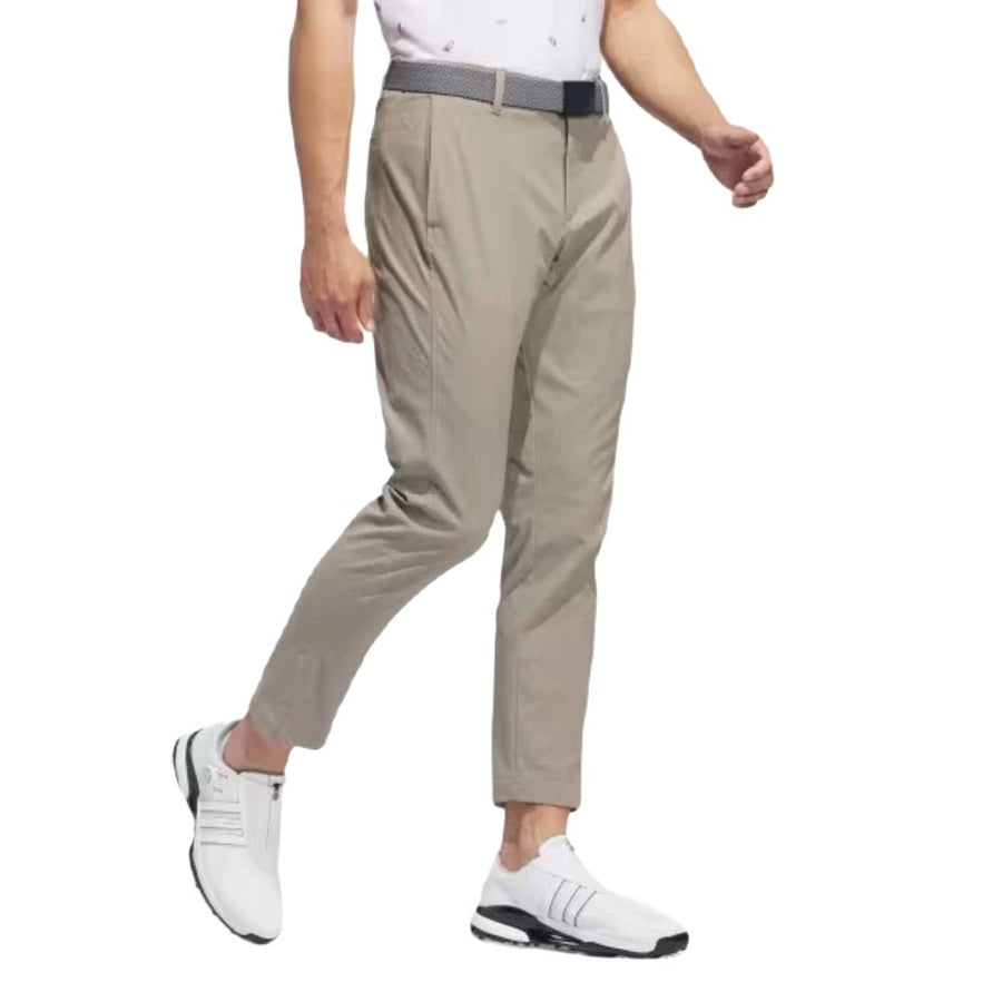 A man wearing light khaki Adidas Ultimate365 Chino Pants made with recycled materials, a black belt, and white athletic shoes with black soles walks with his left leg forward against a white background.