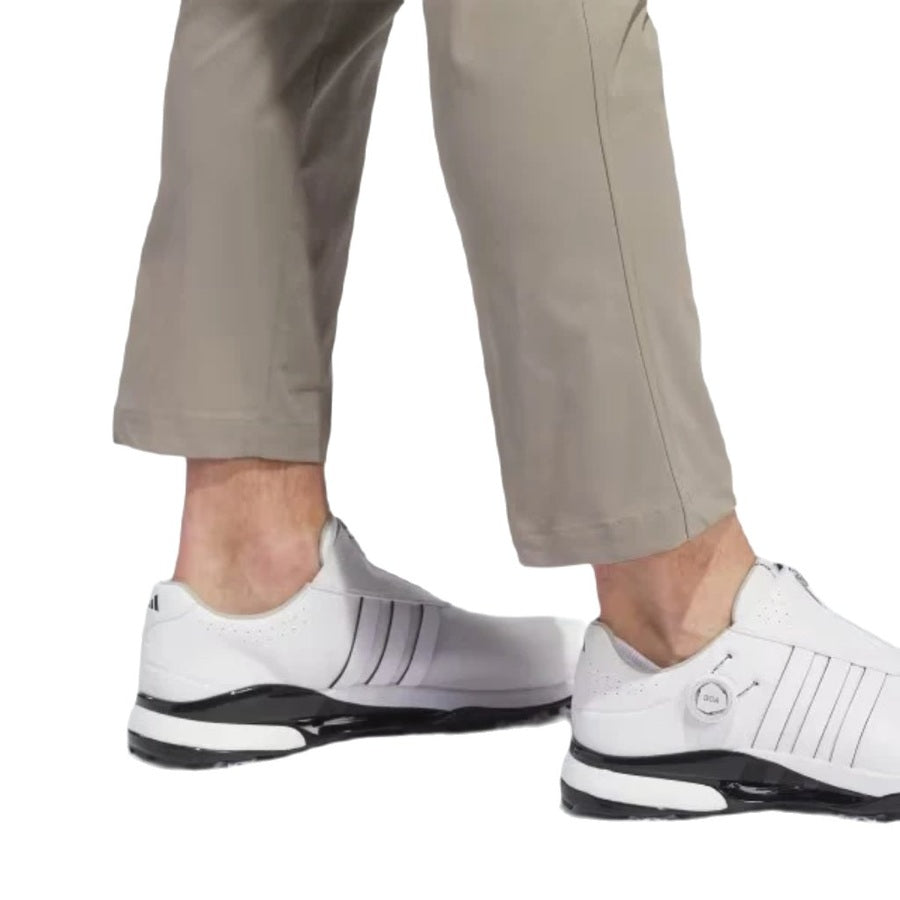 A person is seen from the lower legs down, wearing light khaki Adidas Ultimate365 Chino Pants and white athletic sneakers with black soles, standing against a plain white background.