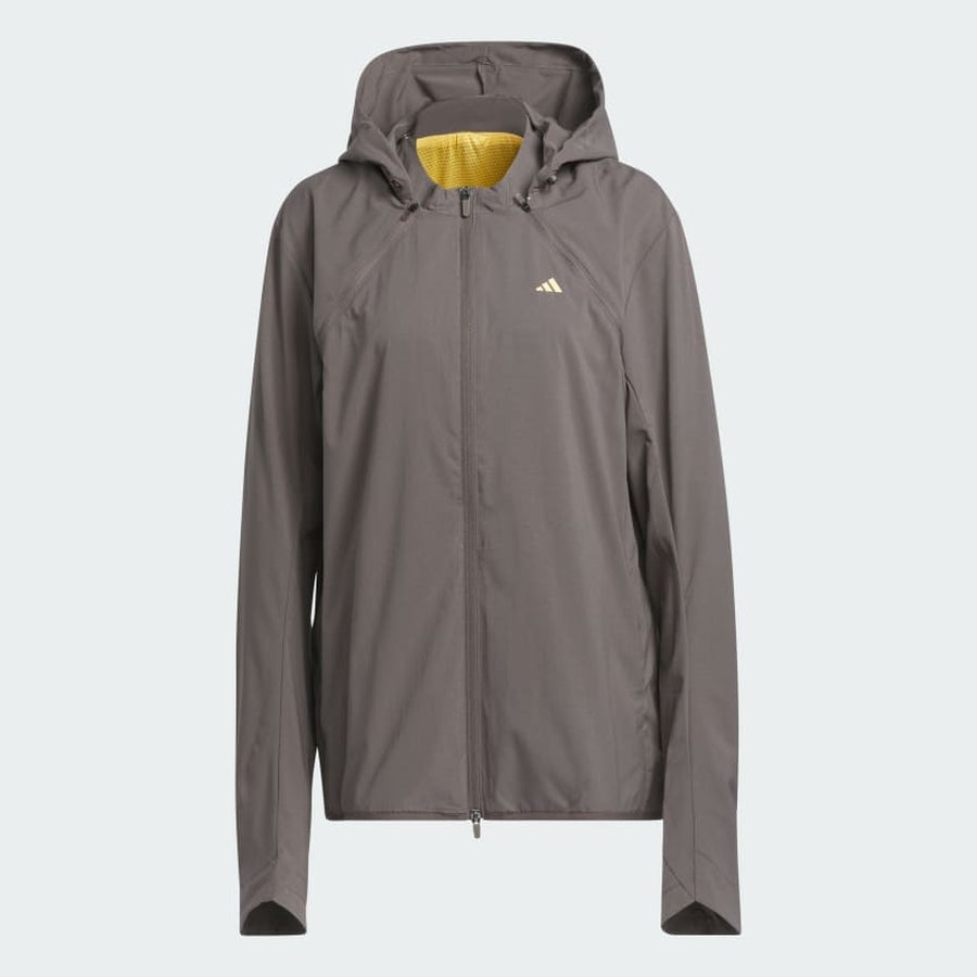 The Adidas Ultimate365 WIND.RDY Convertible Jacket in brown features a hood, long sleeves, front zipper, and small yellow Adidas logo on the left chest. Made with recycled materials, it has a yellow hood lining and is shown on a plain light background.