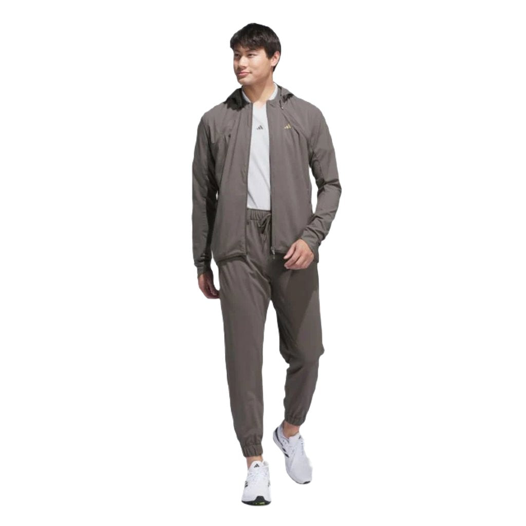 A man in a gray tracksuit and Adidas Ultimate365 WIND.RDY Convertible Jacket - Brown, paired with a white T-shirt and sneakers, walks confidently while looking to the side against a plain white background.