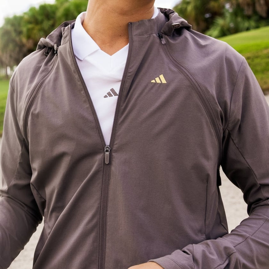 A person wearing a brown Adidas Ultimate365 WIND.RDY Convertible Jacket made with recycled materials over a white Adidas shirt, standing outdoors with greenery and palm trees in the background.
