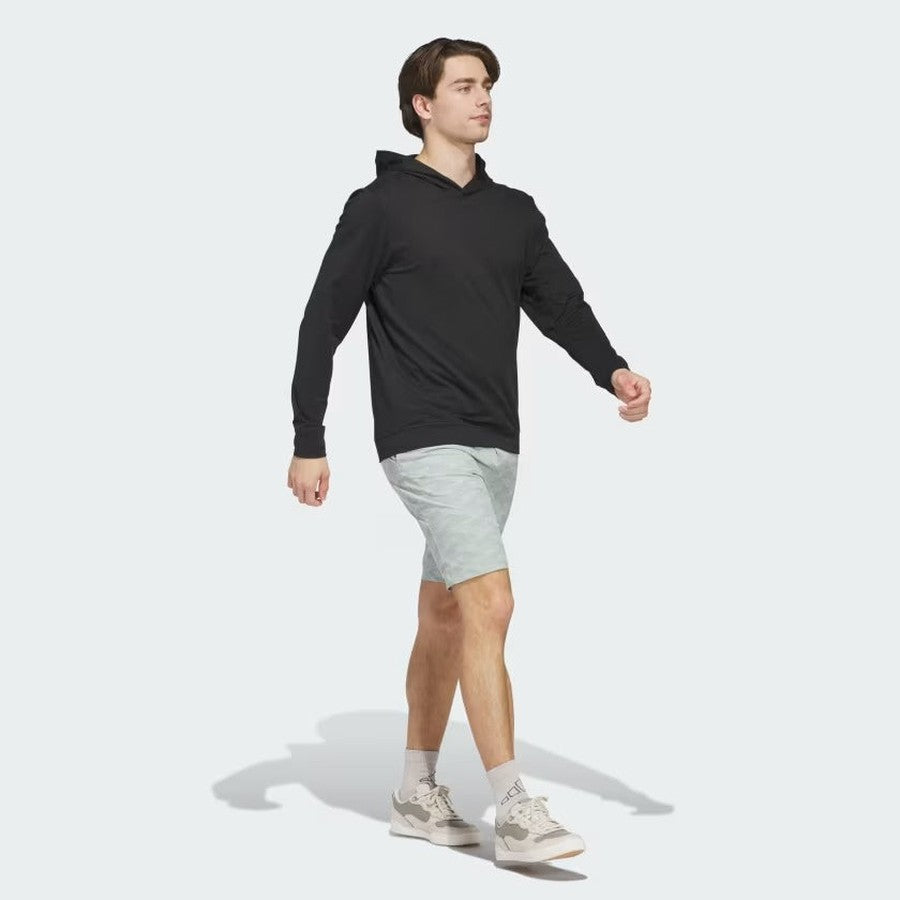 A young man walks confidently in a black Adidas Ultimate365 Elevated Pullover Hoodie made with recycled materials, paired with light gray shorts, white socks, and white sneakers against a plain light gray background.