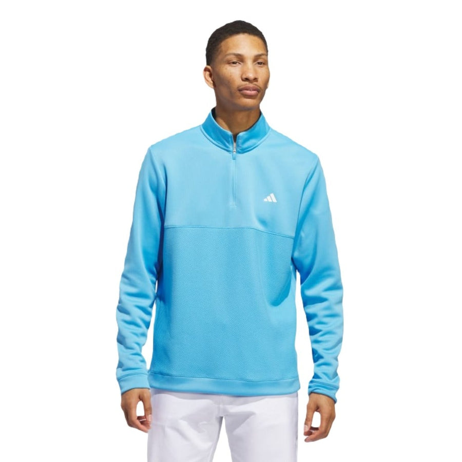 A man wears the Adidas Ultimate365 Textured Quarter-Zip Top—a light blue, long-sleeve pullover with a white Adidas chest logo—paired with white pants, standing against a plain white background.