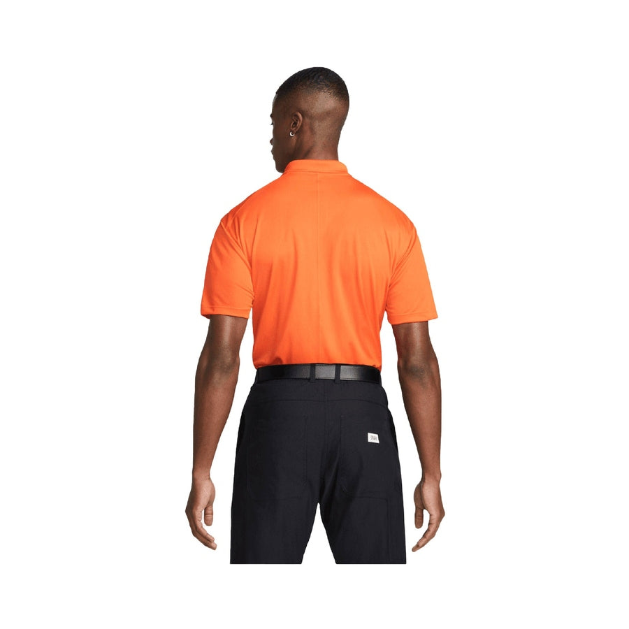 A man faces away, wearing a Nike Dri-FIT Victory Men's Golf Shirt with course/event logo in orange, tucked into black pants with a black belt, against a plain white background.