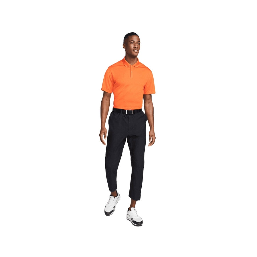 A man stands smiling against a plain white background, wearing a bright orange Nike Dri-FIT Victory Men's Golf Shirt with a course/event logo and "Final Sale" tag, paired with black pants, a black belt, and white sneakers.