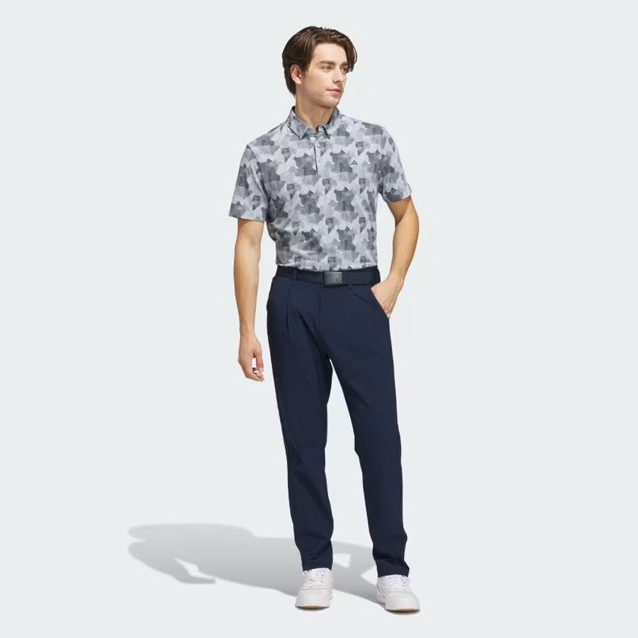 A young man stands with one hand in his pocket, wearing the Adidas 2025 Go-To Printed Polo Shirt in gray camo, navy blue pants, a black belt, and white sneakers against a plain light background.