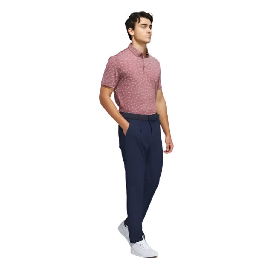 A man wearing an Adidas Ultimate365 Novelty Polo Shirt in burgundy, navy blue trousers, and white sneakers stands with his hands in his pockets against a plain white background.