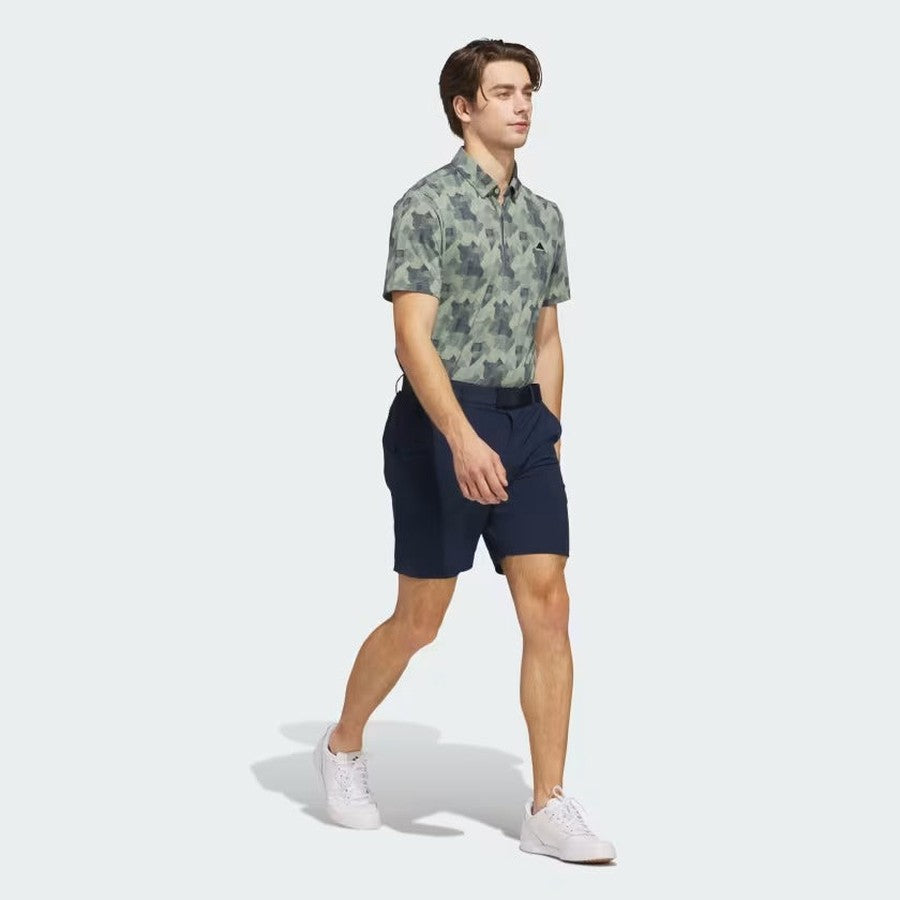 A man walks forward in the Adidas 2025 Go-To Printed Polo Shirt, featuring a short-sleeve green camo print and made from recycled materials, paired with navy shorts and white sneakers, hands in pockets, against a light gray background.