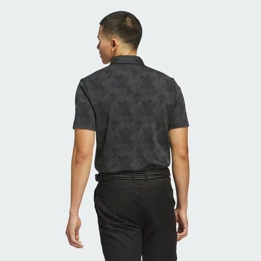 A man with short hair stands facing away from the camera, wearing a dark Adidas 2025 Go-To Printed Polo Shirt, black pants, and a belt, against a plain light background.