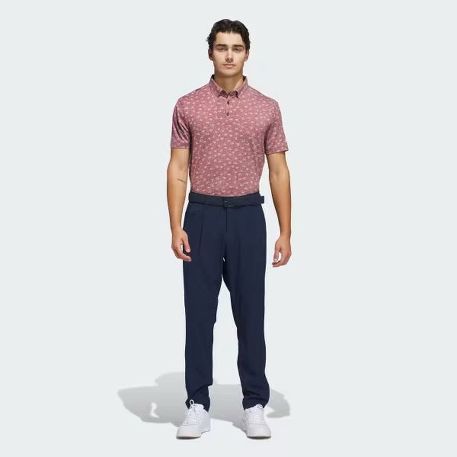 A man faces forward, wearing an Adidas Ultimate365 Novelty Polo Shirt in burgundy, made from recycled materials, paired with navy pants, a black belt, and white sneakers against a plain light background.