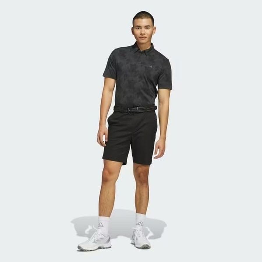 A man with short hair stands against a light background wearing the Adidas 2025 Go-To Printed Polo Shirt made from recycled materials, paired with black shorts, white socks, and white athletic shoes. He looks relaxed with one hand by his side.