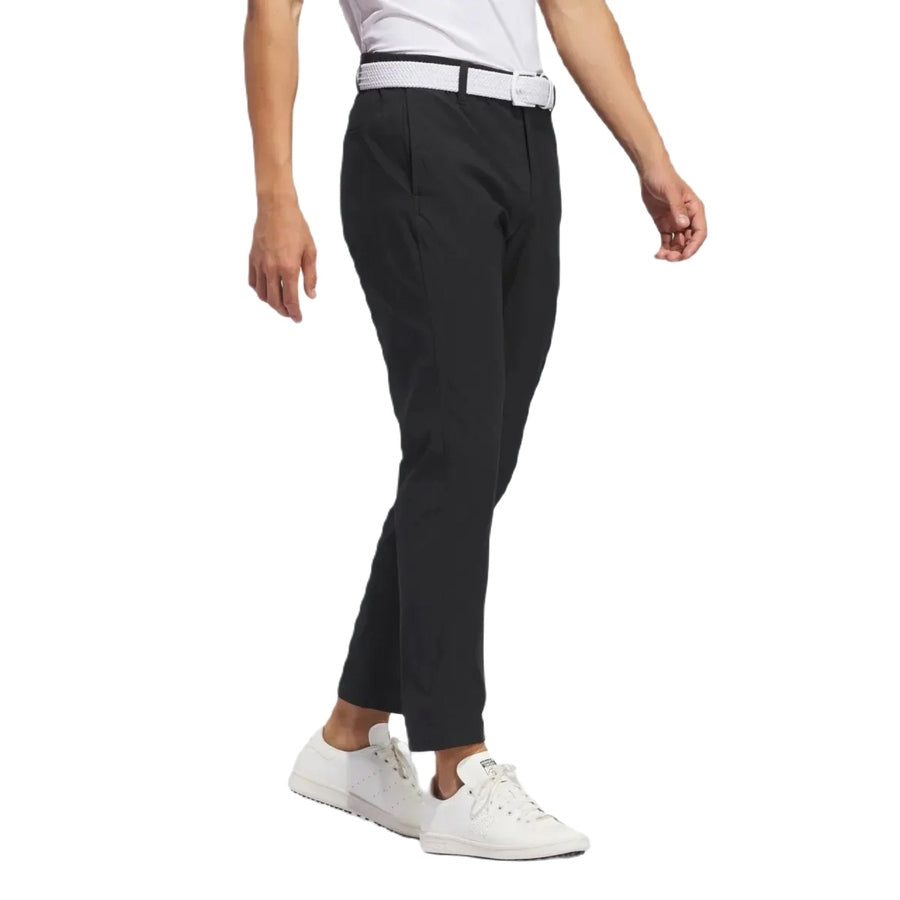 A person is shown walking in black Adidas Ultimate365 Chino Pants made from recycled materials, paired with a white belt and white sneakers. Their upper body is mostly out of frame against a plain white background.