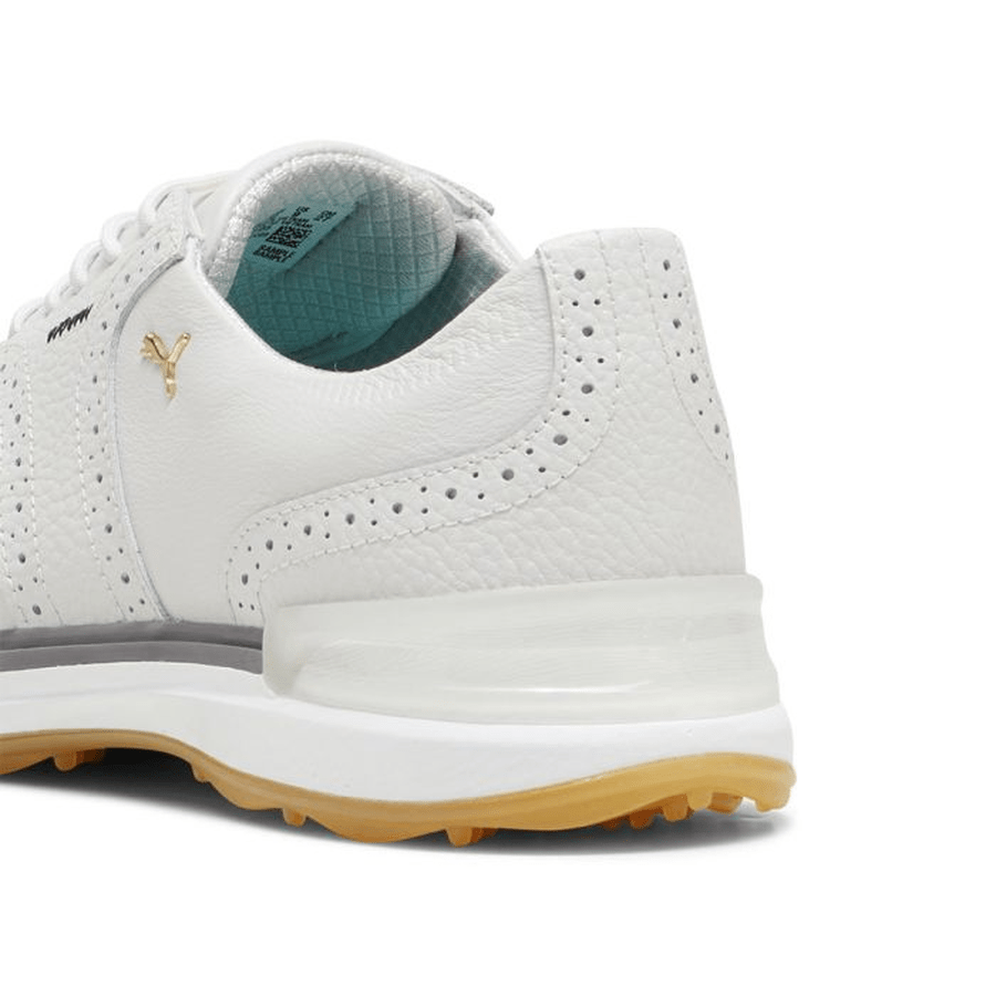 Close-up of the heel and side of the PUMA Avant Wingtip Golf Shoes - Grey, showing a textured white leather upper, gold logo, perforated details, and a spikeless sole with a light brown outsole.