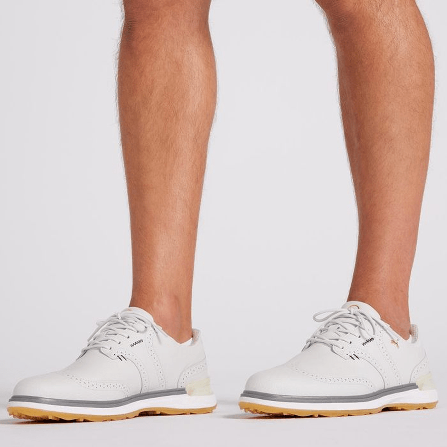 A person wearing PUMA Avant Wingtip Golf Shoes - Grey stands on a plain white background. Only the lower legs and feet are visible, highlighting these stylish grey spikeless golf shoes with light brown and white soles.