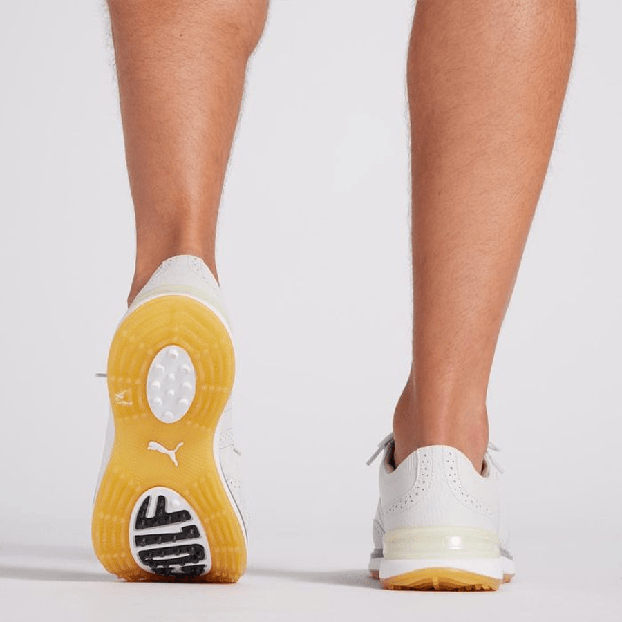 A person is seen from behind wearing PUMA Avant Wingtip Golf Shoes - Grey, showcasing yellow and black detailing on the soles. Their calves are visible against a plain light gray background.