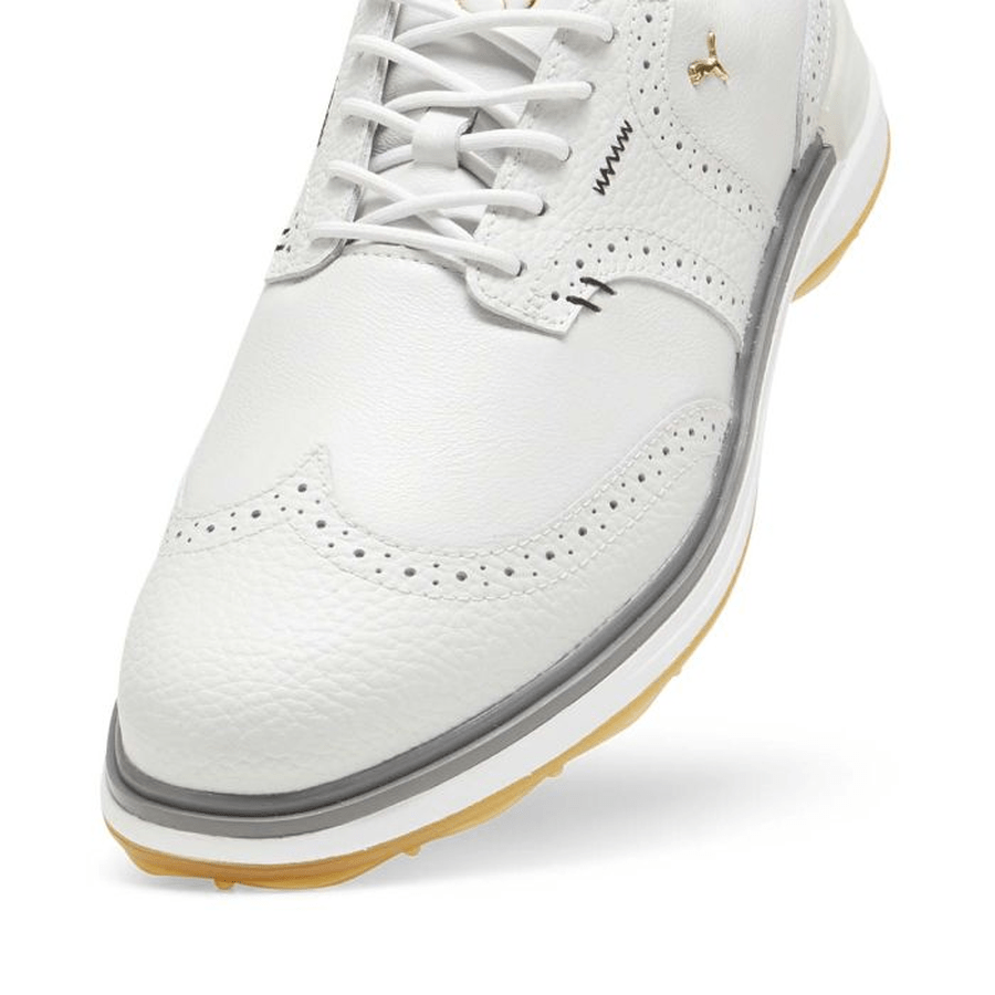 Close-up of PUMA Avant Wingtip Golf Shoes - Grey, featuring grey leather with decorative perforations, white laces, tan rubber sole, spikeless design, detailed stitching, and a small metallic logo on the side.
