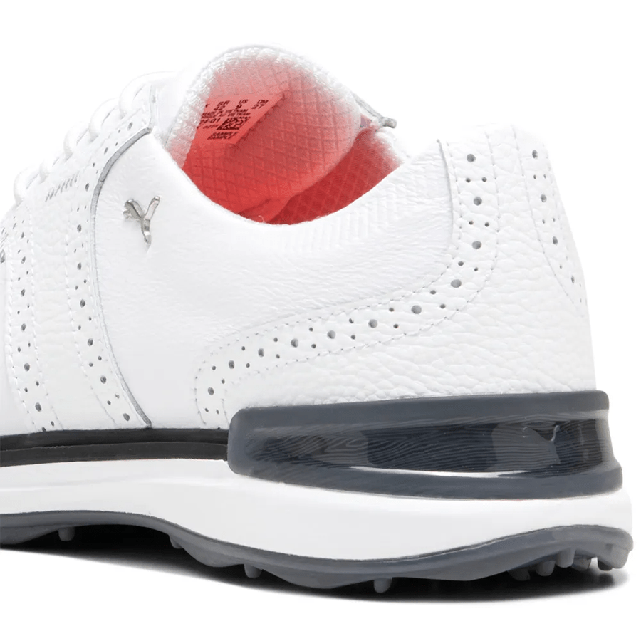 Close-up of the heel and side of the PUMA Avant Wingtip Golf Shoes in white, featuring textured leather, perforated details, radial traction tech, gray heel accent, small Puma logo, gray and white sole, and a red inner lining.